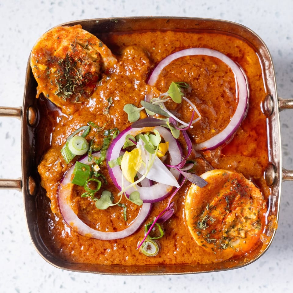 Overhead view of creamy orange curry in a metal serving dish, garnished with grilled seafood, red onion rings, scallions, and microgreens.