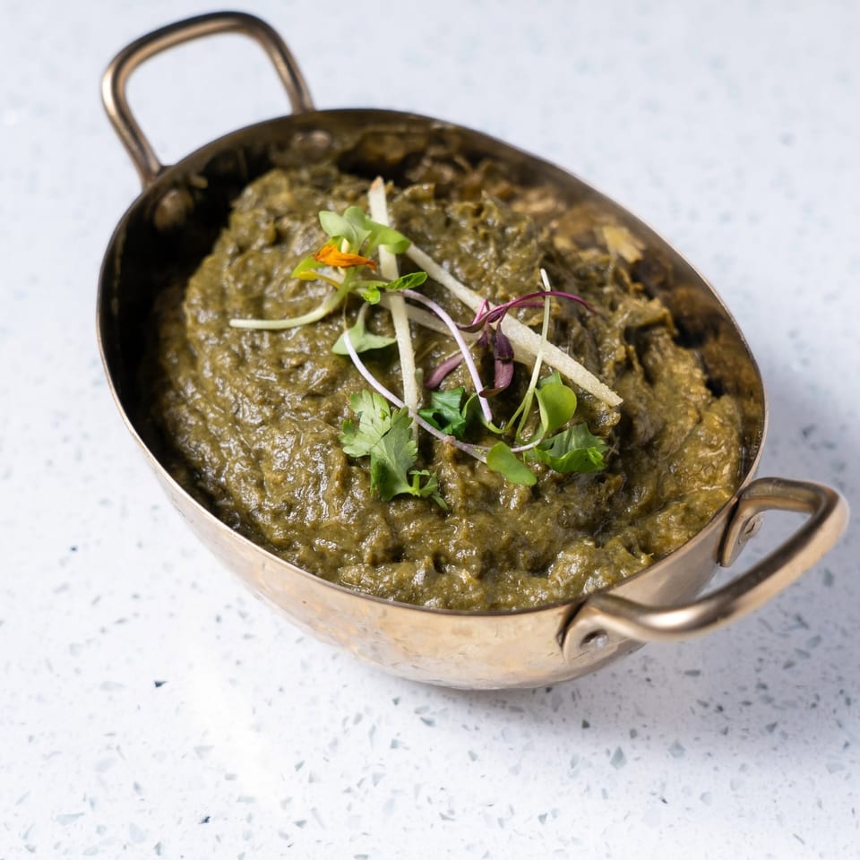 Thick green curry garnished with microgreens and herbs, served in a small brass pan on a light countertop.