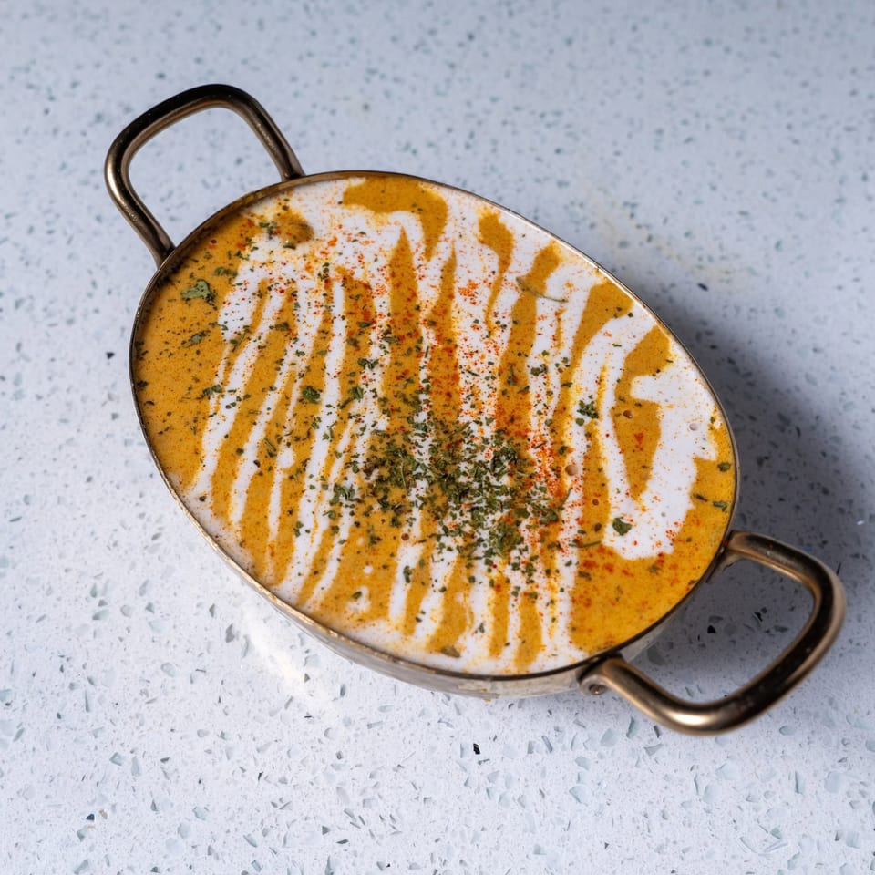 Oval serving dish filled with creamy orange curry, drizzled with white sauce and garnished with herbs and spices.